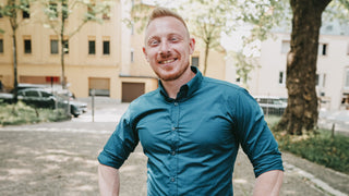 A smiling man in a blue shirt stands in a sunlit tree lined park
