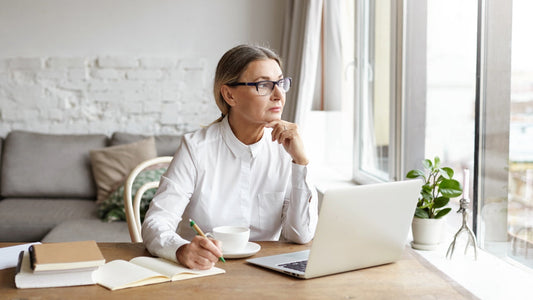 Best Nootropics for Writers in 2025. A woman sits looking out the window pensively while writing on a laptop and a notepad.